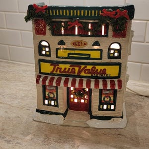 May include: A ceramic True Value Hardware store building for a Christmas village display. The structure is cream-colored with a red and white striped awning, green trim, and festive wreaths. It features the text True Value Hardware and 1948.