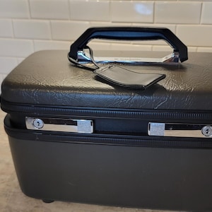 May include: A rectangular, dark gray hardshell vanity case with a textured top surface, a black plastic handle, and two silver-toned metal latches on the front. A small, matching dark gray luggage tag is attached to the handle base.
