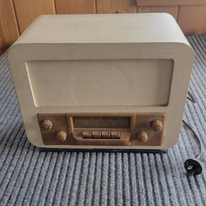 May include: A cream-coloured vintage radio with a rectangular speaker grille and brown control panel. The radio has rounded edges and a power cord. The controls include knobs and buttons. It sits on a grey, ribbed surface.