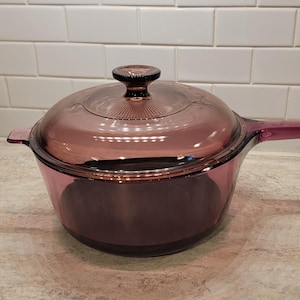 May include: A translucent purple glass saucepan with a matching glass lid. The cookware features a round body, a single long handle on one side, and a small tab handle on the opposite side. The lid has a central knob handle for easy removal.