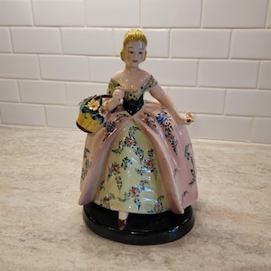 May include: A vintage porcelain figurine of a woman in a long gown, holding a basket of flowers. The dress is pink with floral patterns, and the base is black. The woman has blonde hair and is wearing a green and black bodice.