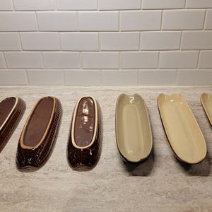 May include: Six ceramic corn-shaped serving dishes. Three are brown with a white rim and three are beige. The dishes are arranged in a row on a light-coloured countertop, with a white tile backsplash.