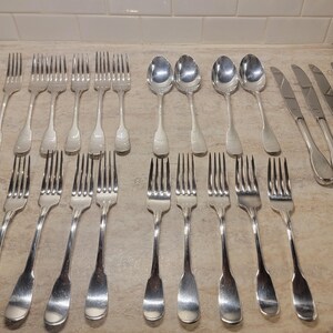 May include: A collection of vintage silverware, including forks, spoons, and knives, arranged on a light-colored surface. The silverware is made of a shiny, silver-colored metal. The set includes various sizes of forks, spoons, and knives, suitable for a formal dining setting.