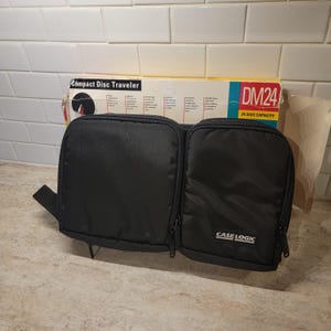 May include: A black nylon Case Logic Compact Disc Traveler storage case with two zippered compartments and a 24 disc capacity. The product is displayed in front of its original cardboard packaging featuring the text Compact Disc Traveler DM24.