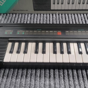 Puede incluir: Un teclado electrónico Yamaha negro con teclas blancas y negras. El teclado tiene una serie de botones y luces encima de las teclas. El teclado está sobre una superficie gris, con una caja en el fondo.