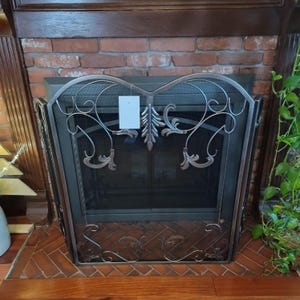 Puede incluir: Una pantalla para chimenea de metal en tono bronce oscuro con un motivo central de hoja vertical flanqueado por curvas simétricas. La pantalla tiene una malla negra fina y una etiqueta blanca rectangular sujeta al marco superior.