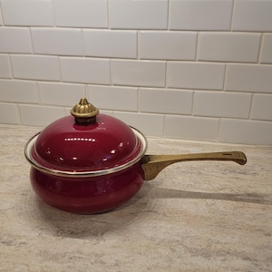 May include: A burgundy saucepan with a lid and a brass handle. The pan has a white rim and a brass knob on the lid. The handle is also brass. The pan is on a speckled countertop.