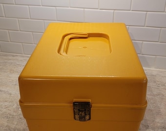 Puede incluir: Una caja de almacenamiento vintage de color amarillo mostaza con una superficie texturizada. La caja tiene forma rectangular con un asa en la parte superior y un cierre de cierre metálico en la parte delantera. La caja está sobre una encimera.