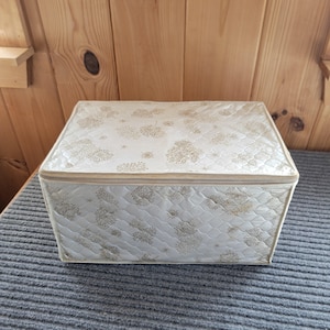 May include: A rectangular storage box with a zippered lid, featuring a quilted white fabric exterior adorned with a repeating gold dandelion and star pattern. The container has structured sides and a flat top, providing a clean, organized look.