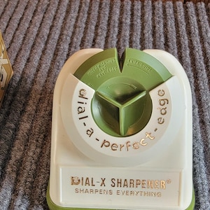 May include: A vintage Dial-X sharpener with a white and green plastic body. The text "Dial-X Sharpener" and "Sharpens Everything" are visible. The sharpener has a green rotating section with "Hollow Ground and Wave Edge" and "Knife Guide" text.