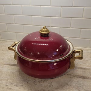May include: A burgundy-colored cooking pot with a matching lid. The pot has gold-colored handles and trim. The lid is topped with a gold-colored finial. The pot is sitting on a light-colored countertop.
