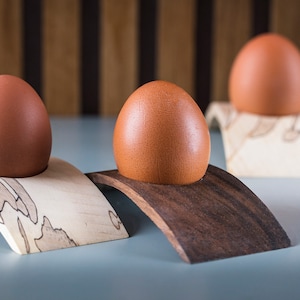 Puede incluir: Tres huevos marrones presentados sobre hueveras de madera. Los soportes son arqueados, con dos de madera clara y uno de madera oscura. El fondo presenta un panel de madera a rayas.