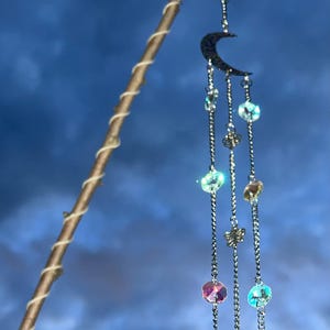 May include: A celestial-themed mobile featuring a silver crescent moon and dangling chains adorned with iridescent beads and charms. The mobile is suspended from a wooden stick wrapped with twine, set against a blue sky background.