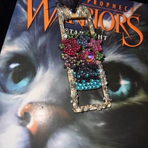 May include: A rectangular bookmark featuring a floral arrangement design embellished with multi-colored rhinestones in shades of pink, green, blue, and purple, finished with a black ribbon at the top and a clear crystal border along the frame.