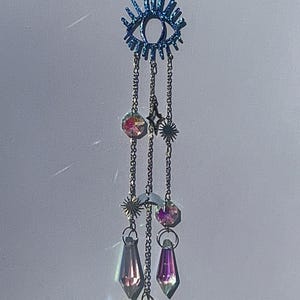 May include: A blue eye-shaped suncatcher with dangling chains. The chains hold iridescent crystals, stars, moons, and heart-shaped pendants. The suncatcher is designed to catch and reflect light.