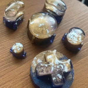 May include: A collection of handmade resin game controller buttons in various shapes. The buttons feature a gold and silver glitter design over a translucent base, with dark blue accents. The buttons are arranged on a wooden surface.