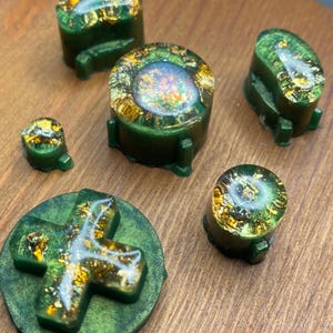 Può includere: Una collezione di pulsanti per controller di gioco in resina verde con accenti di foglia d'oro. Il set include varie forme, come rotonde, ovali e un D-pad. I pulsanti hanno una finitura lucida e sono disposti su una superficie di legno.