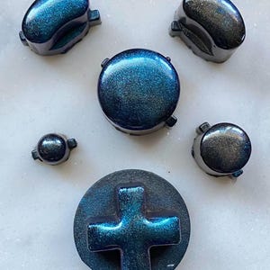 Puede incluir: Un conjunto de botones de mando de videojuegos azul y negro iridiscentes. El conjunto incluye botones ovalados, redondos y en forma de cruz, todos con un acabado brillante y moteado. Los botones están dispuestos sobre una superficie clara.