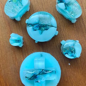 Könnte beinhalten: Eine Sammlung hellblauer Gamecontroller-Tasten aus Harz. Jede Taste hat eine transparente Harzoberfläche mit eingebetteten getrockneten Blättern und blauen und grünen Akzenten. Die Tasten sind auf einer Holzoberfläche angeordnet.