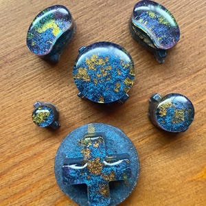 May include: A collection of custom-made video game controller buttons. The buttons are a mix of blue, gold, and purple resin. The D-pad and buttons have a galaxy-like appearance, with gold flecks. The buttons are arranged on a wooden surface.