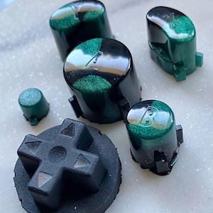 Może przedstawiać: Zestaw niestandardowych przycisków do kontrolera i pad kierunkowy. Przyciski są czarne z zielonym, błyszczącym wierzchem i literami A i B. Pad kierunkowy jest czarny i w kształcie krzyża.