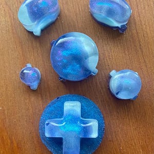 Könnte beinhalten: Eine Sammlung von blauen und violetten Spielcontroller-Tasten aus Kunstharz. Das Set enthält ein Steuerkreuz, runde Tasten und andere geformte Tasten. Die Tasten haben ein glitzerndes, galaxieartiges Aussehen und liegen auf einer Holzoberfläche.