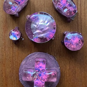 Könnte beinhalten: Eine Sammlung von maßgefertigten, irisierenden lila und rosa Gaming-Controller-Tasten. Das Set enthält ein Steuerkreuz, mehrere ovale Tasten und kleinere runde Tasten, alle mit einem einzigartigen, strukturierten Design.
