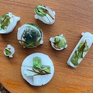 May include: A collection of white resin pieces with embedded green leaves and glitter. The shapes include a circle, a plus sign, a rectangle, and various button-like forms. The objects are arranged on a wooden surface.