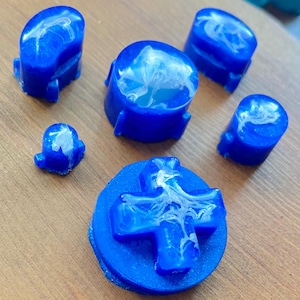 Peut inclure: Ensemble de boutons et d'une croix directionnelle de manette de jeu en résine bleue. Les boutons présentent un motif marbré blanc et bleu. La croix directionnelle est bleue avec un motif marbré blanc, sur une surface en bois.