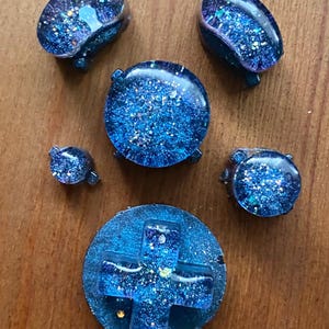 Könnte beinhalten: Eine Sammlung von maßgefertigten Videospiel-Controller-Tasten aus Kunstharz. Die Tasten haben verschiedene Formen und Größen, mit einer durchscheinenden blauen Basis und eingebettetem Glitzer, wodurch ein Sternennachteffekt entsteht. Die Tasten sind auf einer Holzoberfläche angeordnet.