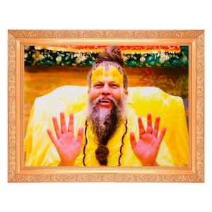 May include: A framed portrait of a man with a long beard and a yellow robe, with his hands raised in a gesture of blessing. He has a yellow mark on his forehead and is smiling.
