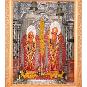 Può includere: Immagine incorniciata con due figure religiose con volti arancioni, copricapi bianchi e abiti rossi e dorati ornati. Sono adornate con ghirlande e poste su uno sfondo argentato dettagliato. La cornice è di un caldo colore dorato.