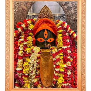 Può includere: Immagine incorniciata di una divinità con volto nero, copricapo arancione e tre occhi. La divinità è adornata da ghirlande colorate di fiori rossi, gialli e bianchi. Un vaso color oro è di fronte alla divinità.