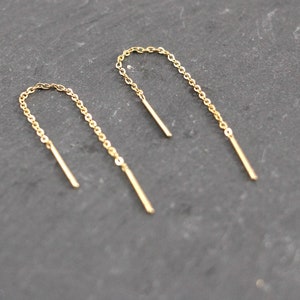 Könnte beinhalten: Ein Paar goldene Kettenohrringe mit schlichtem Design. Die Ohrringe bestehen aus einer dünnen Kette, die oben zu einem kleinen Kreis geschlungen ist. Die Kette hängt dann bis zu einer geraden, dünnen Stange herunter.