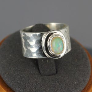 May include: A silver ring with a hammered texture and an oval opal gemstone. The ring is displayed on a wooden stand with a dark gray surface, against a gray background. The opal has a pale green and white color.