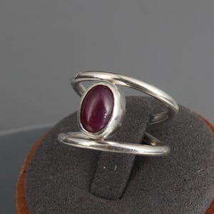 Peut inclure: Bague en argent avec une grosse pierre ovale rouge foncé. La bague a un design moderne et ouvert avec une bande qui entoure la pierre. La bague est présentée sur un présentoir à bijoux en bois.