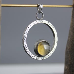 Op de afbeelding: Zilveren hanger met een getextureerd, open cirkelontwerp. Een geel getinte, ronde steen is in de cirkel gezet. De hanger hangt aan een zilveren lus. De achtergrond is neutraal grijs.