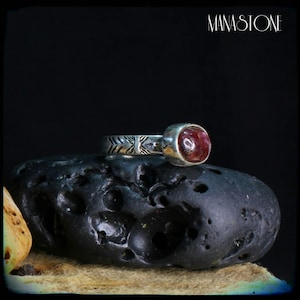 Peut inclure: Une bague en argent avec une pierre précieuse rose foncé sertie dans un sertissage à bélière. La bande de la bague est décorée d'un motif géométrique. La bague repose sur une roche gris foncé.