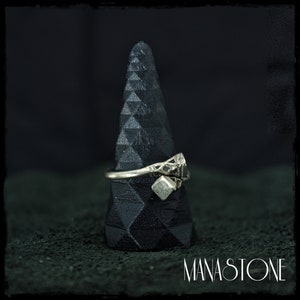 Puede incluir: Un anillo de plata con un diseño geométrico y una pequeña piedra cuadrada. El anillo se muestra en un soporte cónico negro.