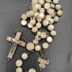 Peut inclure: Un chapelet composé de perles rondes blanc irisé enfilées sur une chaîne en métal sombre. L'objet comprend une pièce de jonction centrale en métal représentant une figure et un crucifix avec une figurine en métal détaillée de Jésus.
