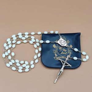 Op de afbeelding: Een witte rozenkrans met ovale kralen en een zilverkleurige metalen ketting en kruisbeeld. De rozenkrans ligt naast een marineblauwe etui met een wapenontwerp en de tekst "INILLO UNO U".