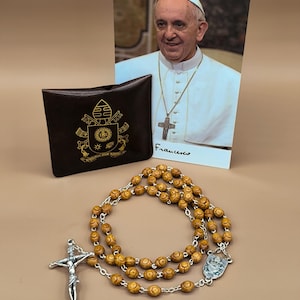 Puede incluir: Un rosario con cuentas de madera y detalles plateados, un pequeño estuche marrón con un emblema dorado y una fotografía del Papa Francisco. El rosario incluye un crucifijo y una medalla.