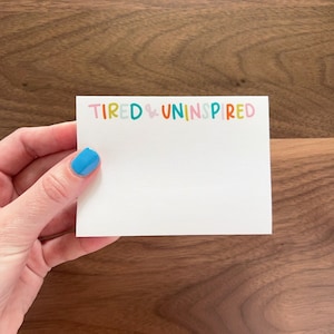 Puede incluir: Una tarjeta blanca con el texto "TIRED & UNINSPIRED" en letras de colores. Las letras están en un estilo juguetón y manuscrito.