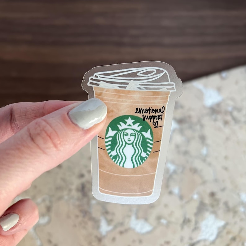May include: A clear sticker of a brown iced coffee cup with a green Starbucks logo and the text 'emotional support' written on the cup.