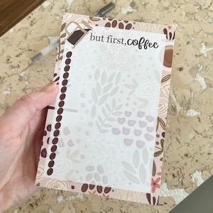 May include: A white notepad with a floral design border. The notepad has a brown coffee pot illustration and the text "but first, coffee" at the top.