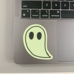 May include: A glow-in-the-dark ghost sticker with black eyes on a gray laptop.