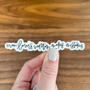 May include: A white sticker with the phrase "even louis vuitton makes mistakes" in black cursive font. The sticker is held by a person with a light-colored nail polish.