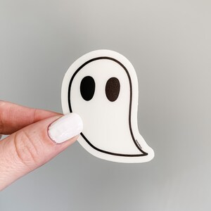 May include: White ghost sticker with black eyes. The sticker is shaped like a ghost with a rounded head and a long, flowing tail.