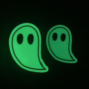 A simple, minimal illustration of a ghost with two black eyes and a black outline. The body of the ghost is white and glows in the dark in green. Two ghosts are shown here to show the two different sizes available