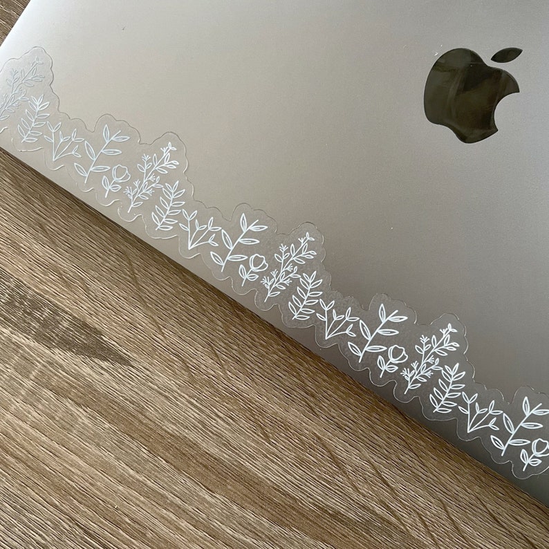 May include: A silver laptop computer with a white floral design sticker on the edge. The laptop is on a wooden surface.